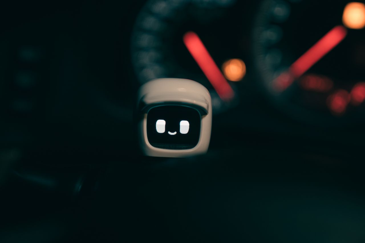 services-02 A small robotic figure with a smiling face on a car dashboard, lit by blurred speedometer lights at night.